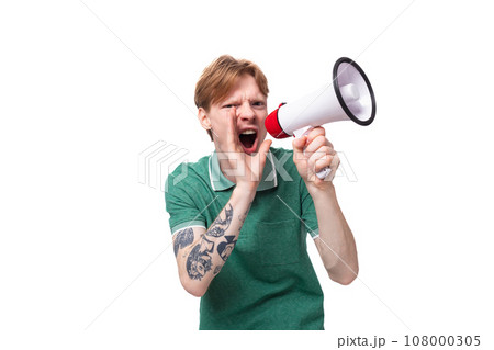 portrait of a surprised red-haired man in a green t-shirt with a tattoo on his arm yells news into a portrait of a surprised red-haired man in a green t-shirt with a tattoo on his arm yells news into a 108000305