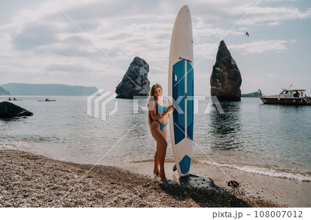 Woman sea sup. Close up portrait of happy young caucasian woman with long hair looking at camera and smiling. Cute woman portrait in bikini posing on sup board in the sea Woman sea sup. Close up portrait of happy young caucasian woman with long hair looking at camera and smiling. Cute woman portrait in bikini posing on sup board in the sea 108007012