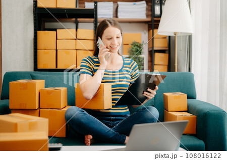 Startup small business SME, Entrepreneur owner woman using smartphone or tablet taking receive and checking online purchase shopping order to preparing pack product box. 108016122
