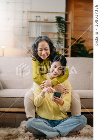 Happy adult granddaughter and senior grandmother having fun enjoying talk sit on sofa in living room at home. Happy adult granddaughter and senior grandmother having fun enjoying talk sit on sofa in living room at home. 108017356