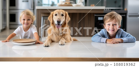Cheerful little boys are posing with a golden retriever dog at the kitchen table. Funny kids and their pet preparing for breakfast at home. Happy smiling brothers and puppy enjoy their time together. 108019362