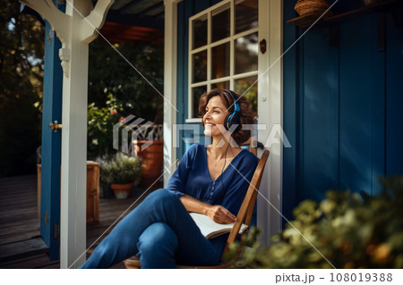 Charming Caucasian woman wearing headphones sits in comfortable chair in a country house. Happy smiling middle-aged lady listens to music and enjoys the silence of her garden. Leisure and relaxation. 108019388