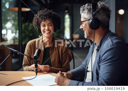 Beautiful multiethnic blogger couple hosting a podcast in a studio. Smiling woman and man wearing headphones sitting at a table with microphones and chatting cheerfully. Modern media technologies. 108019419