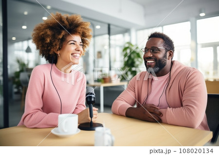 Beautiful multiethnic blogger couple hosting a podcast in a studio. Smiling woman and man wearing headphones sitting at a table with microphones and chatting cheerfully. Modern media technologies. 108020134