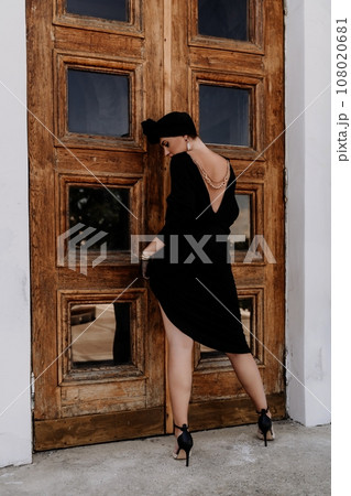 Stylish woman in the city. Fashion photo of a beautiful model in an elegant black dress posing against the backdrop of a building on a city street 108020681
