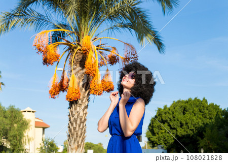 Woman with lush black curly hair in a blue dress near a palm tree and the sky Woman with lush black curly hair in a blue dress near a palm tree and the sky 108021828