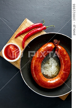 Spiral pork sausage fried in a frying pan with boiling oil on a wooden cutting board with tomato sauce and hot peppers on a black concrete table Spiral pork sausage fried in a frying pan with boiling oil on a wooden cutting board with tomato sauce and hot peppers on a black concrete table 108022866