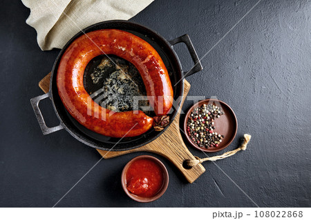 Spiral pork sausage fried in a frying pan with boiling oil on a wooden cutting board with tomato sauce and peppercorns on a black concrete table Spiral pork sausage fried in a frying pan with boiling oil on a wooden cutting board with tomato sauce and peppercorns on a black concrete table 108022868