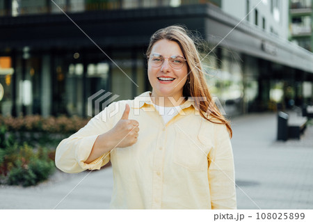 Front view portrait of a happy woman in yellow shirt gesturing thumbs up on the street. Attractive young student girl freelancer looking at camera, posing for photo. Front view portrait of a happy woman in yellow shirt gesturing thumbs up on the street. Attractive young student girl freelancer looking at camera, posing for photo. 108025899