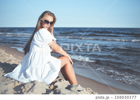 Happy, beautiful blonde woman is sitting on the ocean beach in a white summer dress and sunglasses Happy, beautiful blonde woman is sitting on the ocean beach in a white summer dress and sunglasses 108030377