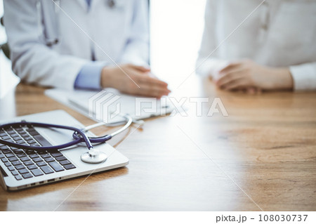 Stethoscope and laptop computer are lying on the wooden table while doctor and patient sitting and talking to each other at the background. Medicine concept Stethoscope and laptop computer are lying on the wooden table while doctor and patient sitting and talking to each other at the background. Medicine concept 108030737