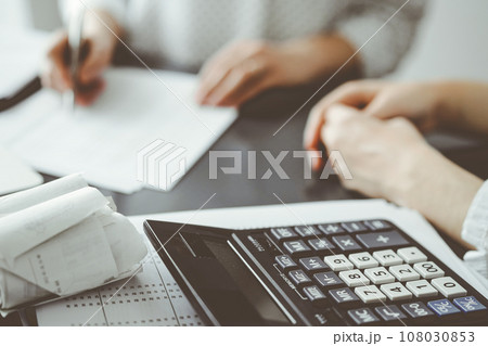 Woman accountant using a pen and laptop computer while counting and discussing taxes with a client, focus on the calculator. Business audit and finance concepts Woman accountant using a pen and laptop computer while counting and discussing taxes with a client, focus on the calculator. Business audit and finance concepts 108030853