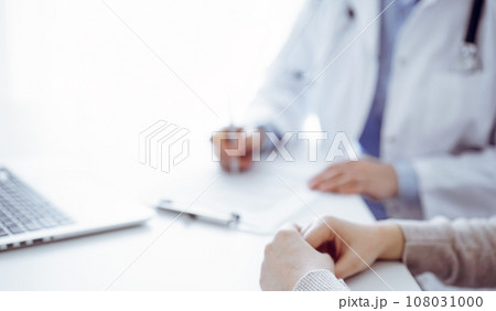 Doctor and patient sitting at the table in clinic. The focus is on female physician's hands filling up the medication history record form or checklist, close up. Medicine concept 108031000