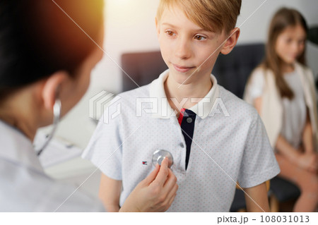 Doctor and kid patient are in the clinic. Physician in white coat examining a smiling young boy with a stethoscope, close up. Medicine, therapy concept 108031013