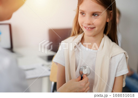 Doctor and kid patient are in the clinic. Physician in white coat examining a smiling young girl with a stethoscope, close up. Medicine, therapy concept 108031018