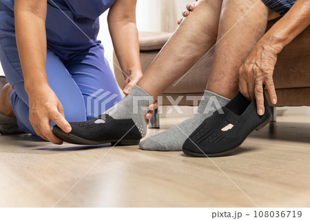 Caregiver helping elderly woman to put shoe at home 108036719