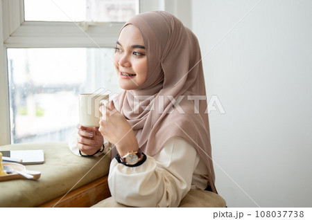 An attractive young Asian woman wearing a hijab is enjoying coffee while relaxing in a cafe alone. 108037738