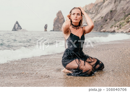 Woman summer travel sea. Happy tourist in black dress enjoy taking picture outdoors for memories. Woman traveler posing on sea beach surrounded by volcanic mountains, sharing travel adventure journey 108040706