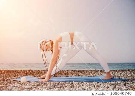 Woman sea yoga. Well looking middle aged woman with braids dreadlocks in white leggings and tops doing stretching pilates on yoga mat near sea. Female fitness yoga routine concept. Healthy lifestyle. Woman sea yoga. Well looking middle aged woman with braids dreadlocks in white leggings and tops doing stretching pilates on yoga mat near sea. Female fitness yoga routine concept. Healthy lifestyle. 108040740