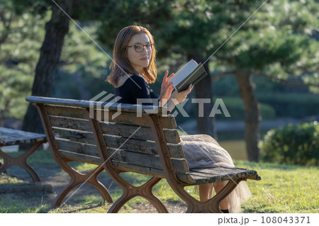 緑に囲まれた公園のベンチで本を読む女性 108043371