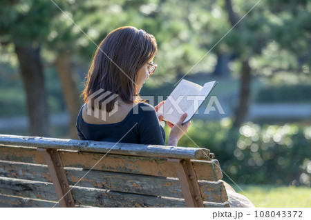 緑に囲まれた公園のベンチで本を読む女性 108043372