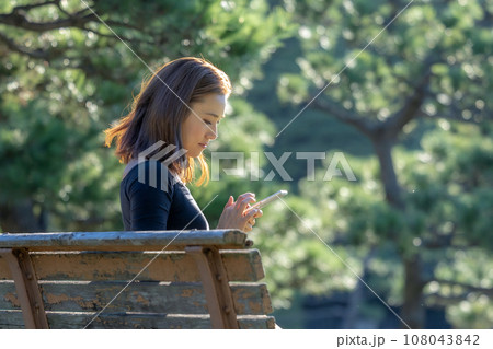 緑に囲まれた公園のベンチでスマホを操作する女性 108043842