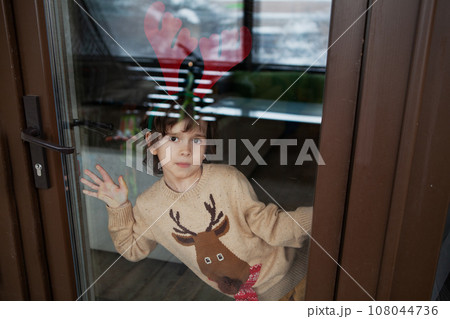 Adorable little boy in knitted beige deer sweater wearing reindeer antlers at home and looking at camera through window glass Adorable little boy in knitted beige deer sweater wearing reindeer antlers at home and looking at camera through window glass 108044736