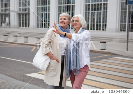 Older women with gray hair in urban landscape, smile, gaze and point off in distance. Fashionable and stylish retired female friends enjoying cityscape and adventure together 108044800
