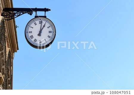 Street clock with Roman numerals on a building and blue clear sky Street clock with Roman numerals on a building and blue clear sky 108047051