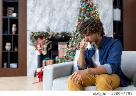 Sick sneezing man with runny nose sitting on sofa at home, hispanic winter day near Christmas tree, cold and allergy in living room inside house. 108047798