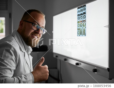 Side view of doctor wearing glasses examining lungs scan. Male specialist wearing white robe, looking at camera, smiling, showing super. Concept of hospital and healthcare. Side view of doctor wearing glasses examining lungs scan. Male specialist wearing white robe, looking at camera, smiling, showing super. Concept of hospital and healthcare. 108053456