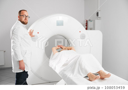 Medical computed tomography or MRI scanner. Male radiologist presses MRI button to examine female patient, looking to the camera. 108053459