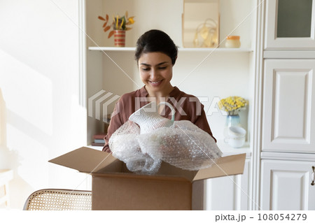 Smiling Indian woman unpacking parcel with fragile order at home Smiling Indian woman unpacking parcel with fragile order at home 108054279