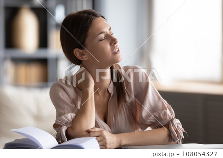 Woman suffers from neck pain sitting at table indoors Woman suffers from neck pain sitting at table indoors 108054327