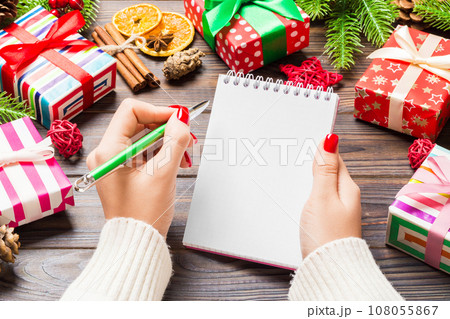 Top view of female hand writing in a notebook on wooden Christmas background. Fir tree and festive decorations. Wish list. New Year concept Top view of female hand writing in a notebook on wooden Christmas background. Fir tree and festive decorations. Wish list. New Year concept 108055867