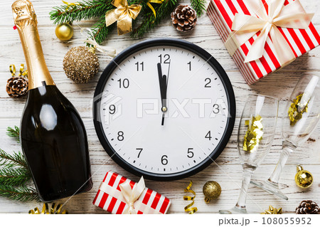New year celebration concept with a bottle of champagne and two glasses toasting. Christmas gift box, alarm clock and fir tree branch on colored table. Top view Copy space 108055952