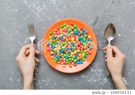 the girl holds cutlery in her hands and eats sweets in a plate. Health and obesity concept, top view on colored background the girl holds cutlery in her hands and eats sweets in a plate. Health and obesity concept, top view on colored background 108056131