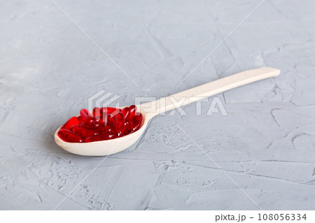 Vitamin capsules in a spoon on a colored background. Pills served as a healthy meal. Red soft gel vitamin supplement capsules on spoon 108056334