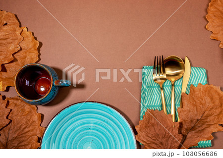 Autumn creative idea of holiday table setting for Halloween or thanksgiving day. Empty turquoise tableware plate and cup, golden cutlery, dry oak leaves on brown background. Top view. Copy space. 108056686
