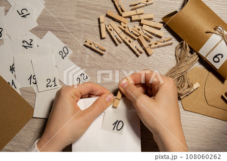 Unrecognizable young woman sticks number on craft bag, fastens with clothespin. Female making kraft paper for homemade advent calendar Made with your own hands step by step DIY crafts do it yourself 108060262