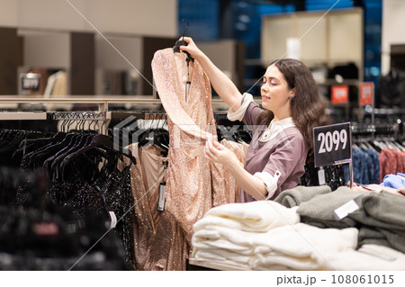 Shopping and sales. Smiling young Caucasian woman holds and looks at hanger with sparkly dress. Concept of consumerism and black friday Shopping and sales. Smiling young Caucasian woman holds and looks at hanger with sparkly dress. Concept of consumerism and black friday 108061015
