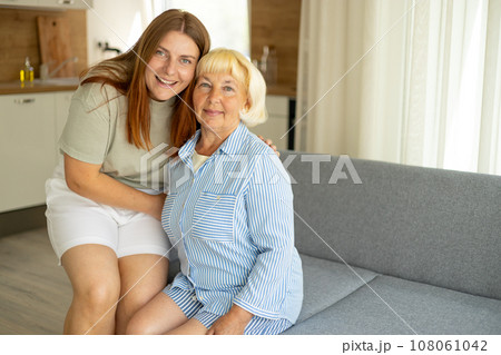 Happy senior woman enjoying in daughter's affection on Mother's day. Smiling mature mother and daughter hugging, sitting on couch at home, looking at camera 108061042