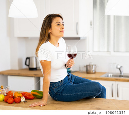 Portrait of relaxed woman sitting on kitchen countertop, drinking red wine 108061355