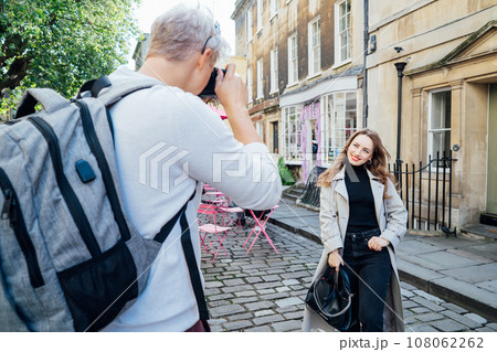 Back view man making photo of smiling young woman outdoors in old European city. Tourists taking picture during travel walks. Professional photo shooting on urban streets. Photographer occupation. 108062262