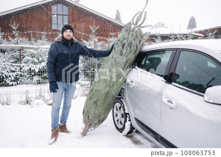 Handsome bearded man stand by the car and think how to put a Christmas tree into car Handsome bearded man stand by the car and think how to put a Christmas tree into car 108063753