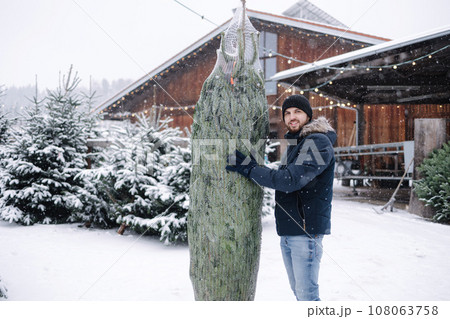 Happy handsome bearded man holds a packed live Christmas tree in outdoor market. Winter snowy day Happy handsome bearded man holds a packed live Christmas tree in outdoor market. Winter snowy day 108063758