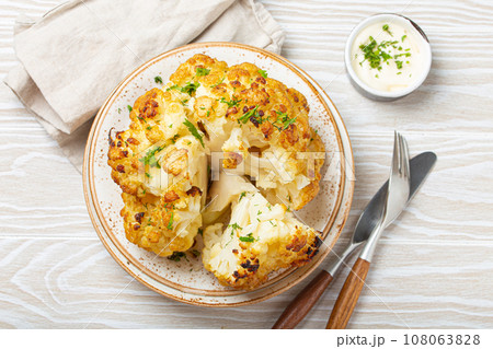 Whole roasted head of cauliflower with olive oil on plate on white wooden rustic table top view with cutlery. Vegetarian dish. 108063828