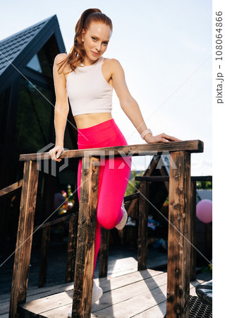 Vertical portrait of sensual athletic young woman in sportswear looking at camera standing leaning on handrail of country house after training, on sunny summer day on background of sky. Vertical portrait of sensual athletic young woman in sportswear looking at camera standing leaning on handrail of country house after training, on sunny summer day on background of sky. 108064866