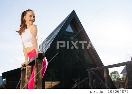 Portrait of positive athletic female in sportswear standing leaning on handrail of country house after training on sunny summer day on background of sky. Sportswoman posing resting outdoors 108064872