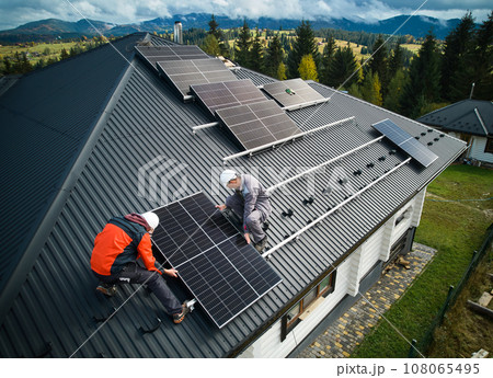 Electricians building photovoltaic solar module station on roof of house. Men technicians in helmets installing solar panel system outdoors. Concept of alternative and renewable energy. Aerial view. 108065495